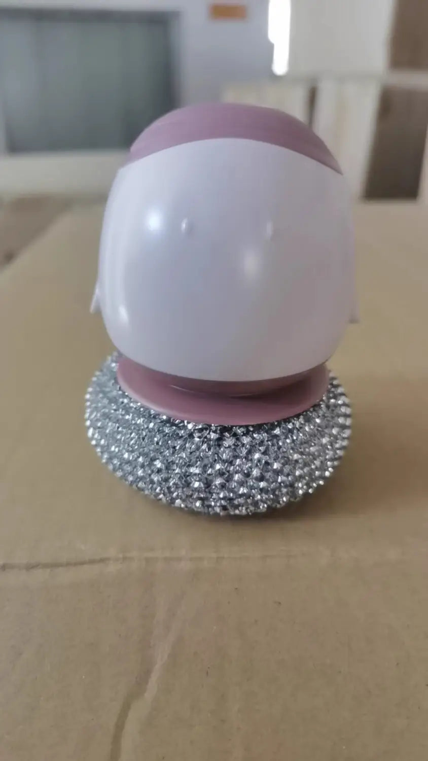 récurant-distributeur automatique de savon liquide pour brosse à plat en forme d’œuf, avec une tête de récurage sphérique rose et blanche et une base argentée scintillante à petits grains.