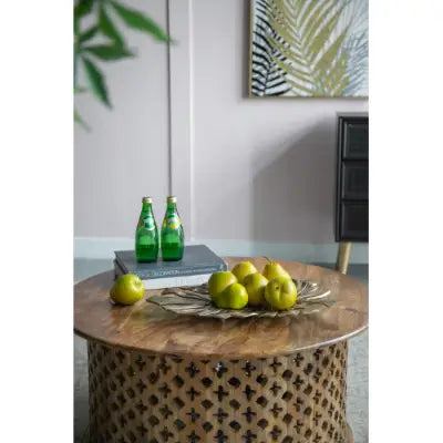 Round wooden coffee table with intricate lattice design and natural wood finish featured alongside gold monstera leaf wall decor and serving tray.