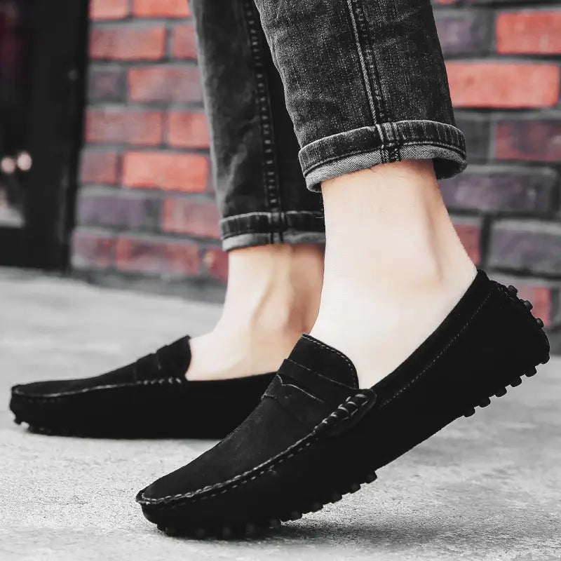 Mocassins en daim pour homme noirs, avec une lanière en cuir cousue sur l’avant et des semelles en caoutchouc texturées.