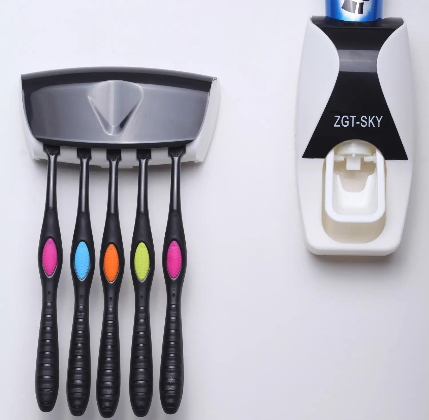 Black toothbrushes with colorful oval accents on the handles are mounted in a sleek gray and white wall-mounted automatic toothpaste dispenser and toothbrush holder set.