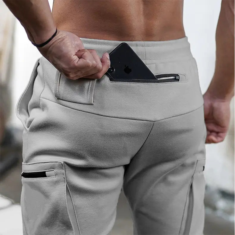 Men’s light gray athletic zipper cargo joggers made of subtle textured fabric with black zipper pockets designed for fitness use.