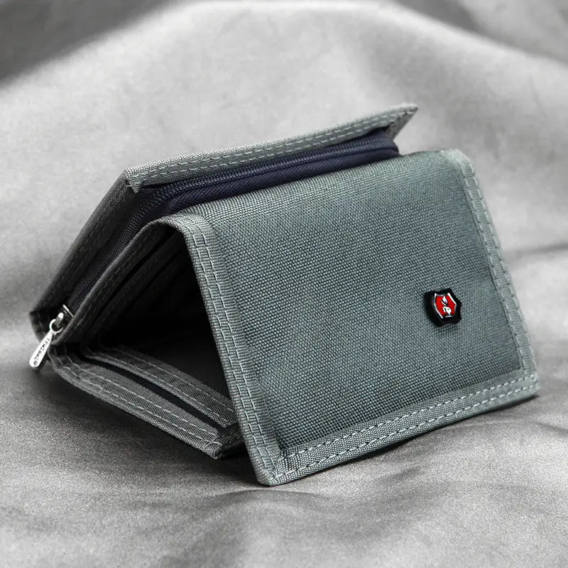 Cartera táctica de lona gris tipo billetera plegable en tres con un logotipo de escudo rojo y negro en el frente, diseñada como portamonedas.