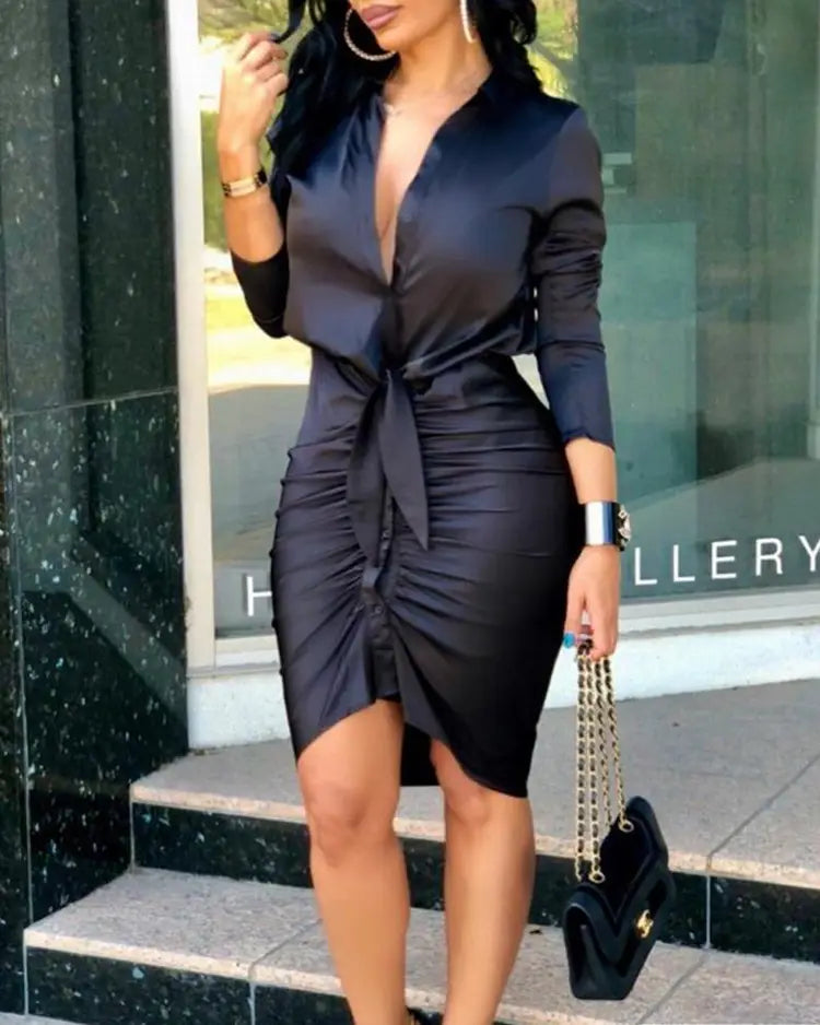 Black satin sheath dress with a deep v-neckline and tied waistband featuring long sleeves and ruched detailing for an elegant midi length.