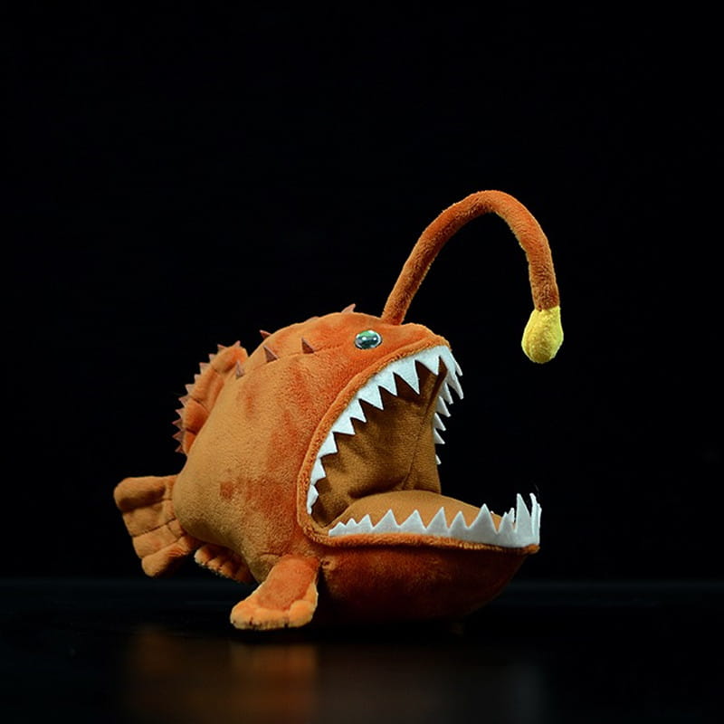 Orange plush anglerfish glow lantern toy with white jagged teeth and a yellow-tipped fishing lure on its curved dorsal fin measuring 25cm.