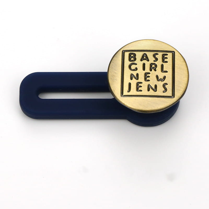 Extendeur de bouton de taille de jeans ajustable sans couture, présentant une pince en silicone bleu marine avec un emblème circulaire de couleur laiton gravé du texte « base girl new jens » dans un cadre carré.