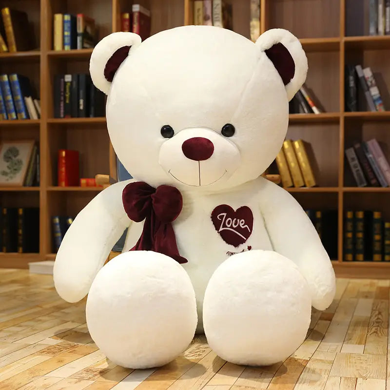 Weiße Plüsch-Liebesbär-Plüschfigur mit Herz, eine Teddy-Bär-Figur mit maroonfarbenen Akzenten an Ohren, Nase und Fliege; mit einem herzförmigen, bestickten Patch mit dem Wort „love“ auf der Brust.