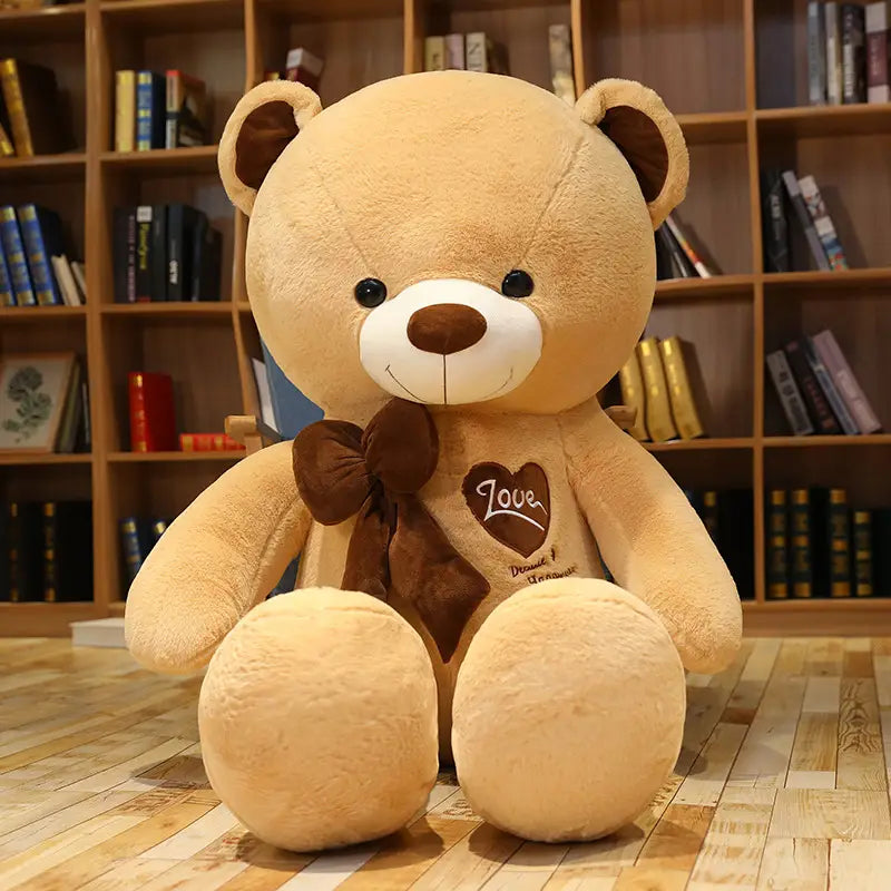 Ein beiger Plüsch-Teddybär mit braunen Akzenten, schwarzen Knopfaugen, einem gestickten Lächeln und einer passenden Fliege; mit dem Wort „love“, bestickt auf seiner Brust, aus der Kollektion „Love Bear Plüschfigur – Herzbeichte-Teddy“.