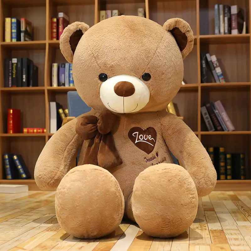 Ein beiger Plüsch-Liebesbär-Plüschspielzeug als Herzbeichte-Teddy mit einem weißen Schnauzenbereich, brauner Nase und Augen, mit einer passenden Schleife um seinen Hals.