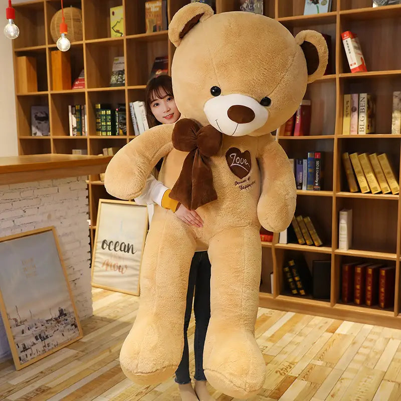 Ein beiger, plüschfarbener Liebesbär-Plüschspielzeug als Herzbeichte-Teddy mit weißem Schnauzenbereich, brauner Nase und besticktem „love“ auf der Brust sowie einer passenden braunen Schleife um den Hals