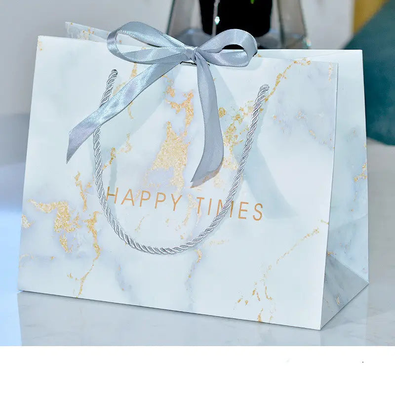 A white and gold marble-patterned paper gift bag with silver satin ribbon bow and twisted silver rope handles displaying happy times is shown alongside a 3d wooden steam locomotive puzzle model kit.