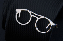 white-framed glasses with round lenses resting on a dark textured surface in a men’s airplane tie clip and lapel pin gift set