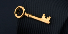 Gold-colored skeleton key pin with a classic design from the men’s airplane tie clip and lapel pin gift set resting on dark fabric.