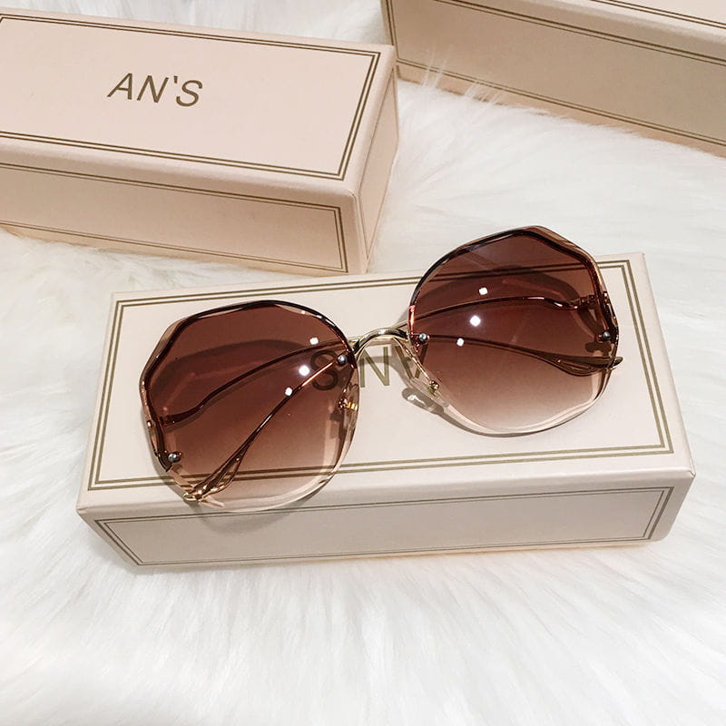 Gradienten Sonnenbrille mit übergroßem, geometrischem Design in Braun, mit goldfarbenem Metallrahmen, die auf einer An’s-Markenbox liegt.