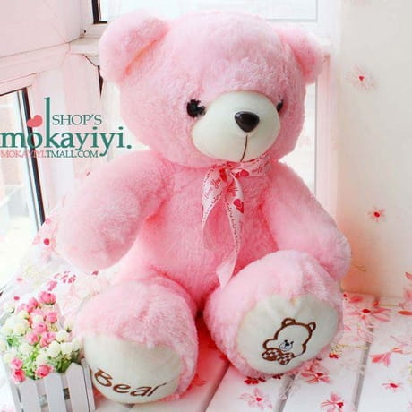 A plush pink teddy bear with a soft texture featuring a white snout, black button eyes, and a delicate ribbon bow around its neck.