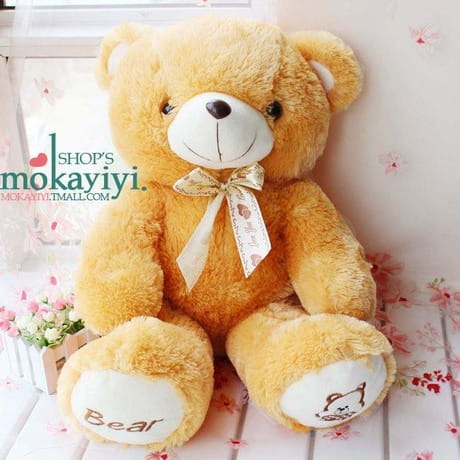 A plush teddy bear soft animal doll in golden brown with a white snout and black button eyes wearing a delicate gold ribbon bow around its neck.