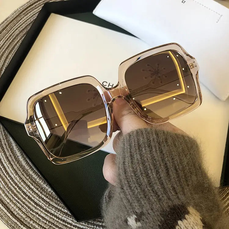 A pair of oversized square fashion sunglasses with translucent amber-colored frames and dark brown mirrored lenses held by a hand wearing a gray knit sweater.