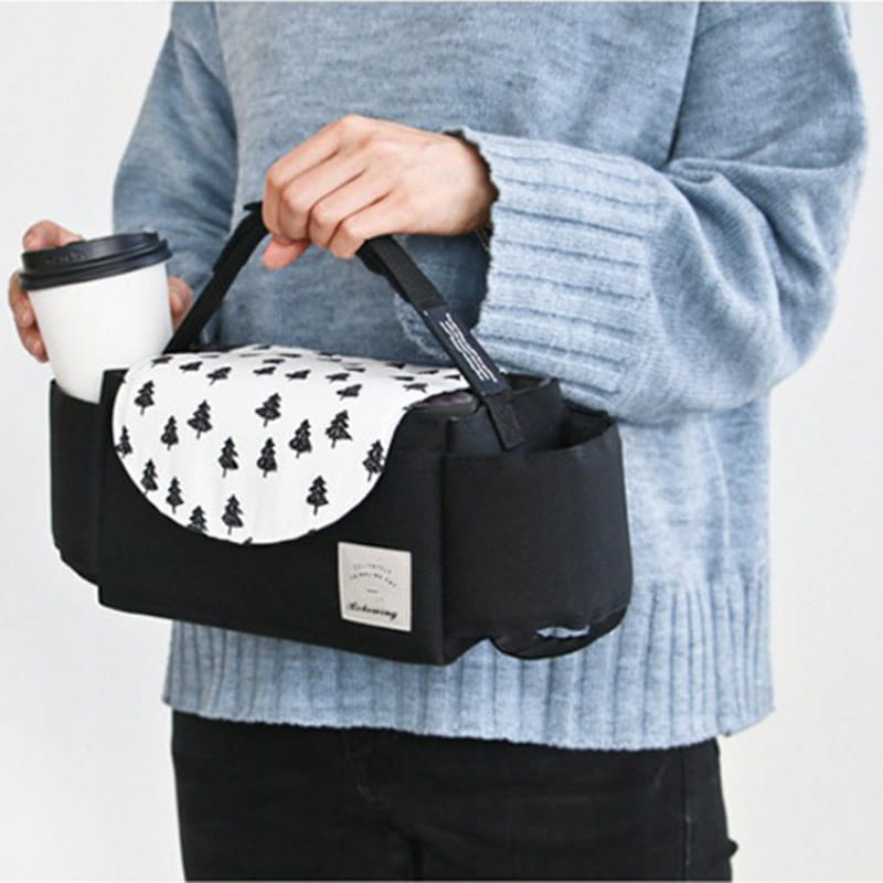 Black fabric coffee cup holder with a white flap featuring black tree patterns and a small rectangular brand label attached to a stroller organizer bag with multi storage pockets.