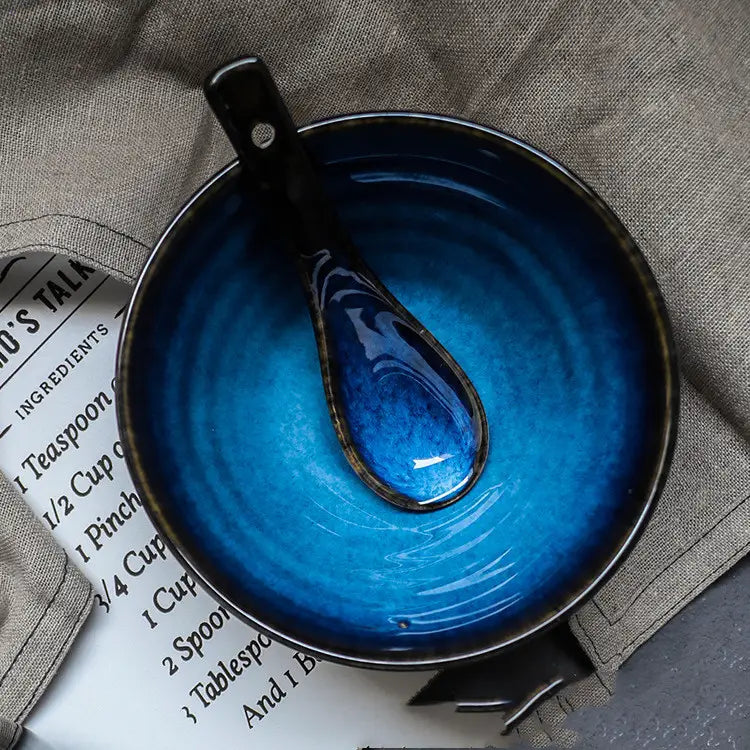 A glossy cobalt blue ceramic bowl with matching hand-painted kiln glaze spoon featuring rustic black rims for porcelain tableware use.