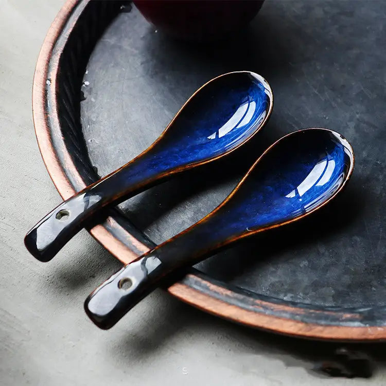 Two glossy blue ceramic spoons with a deep kiln glaze and dark brown handles resting on a rustic copper-rimmed plate in porcelain tableware.