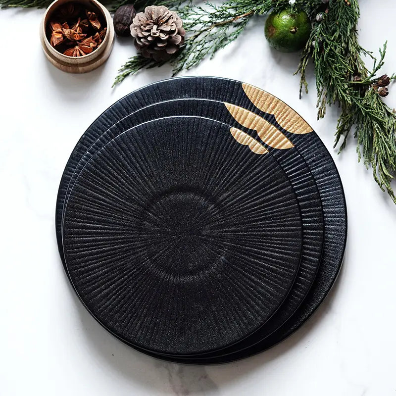 Assiette noire en céramique avec un motif radial texturé, des accents de feuille d’or et un bord en bois.