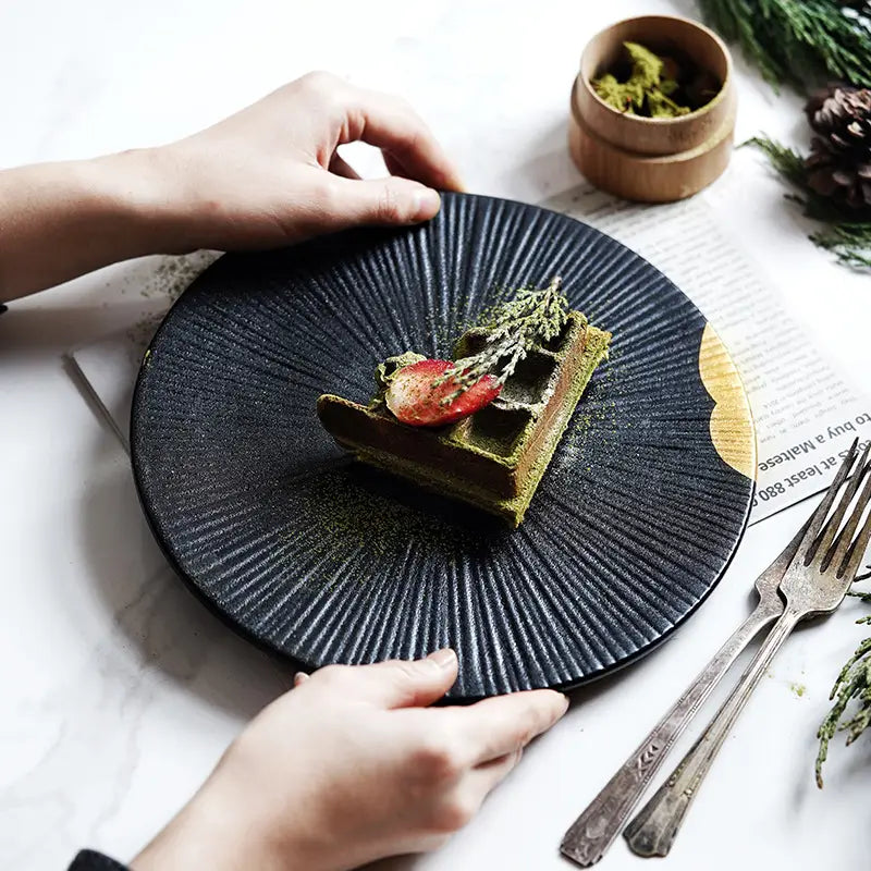Assiette noire en céramique présentant des rainures radiales et une finition mate, avec des accents dorés et d’un bord en bois.