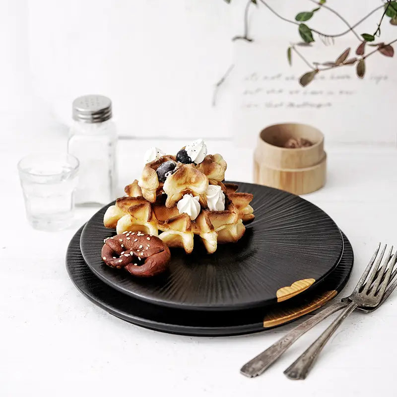 Gaufre brun doré à la texture croustillante, surmontée de crème fouettée et de baies foncées, servie sur une assiette noire en céramique, rehaussée d’accents dorés et d’un bord en bois.