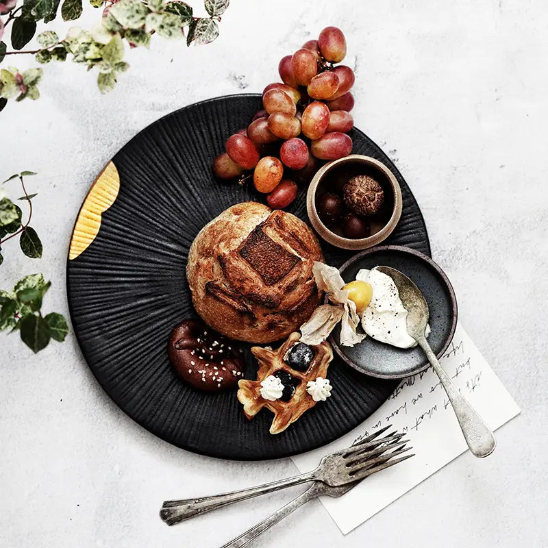 Assiette noire en céramique texturée avec des rainures radiales et un accent doré sur le bord, mettant en valeur des accents élégants de bord en bois.