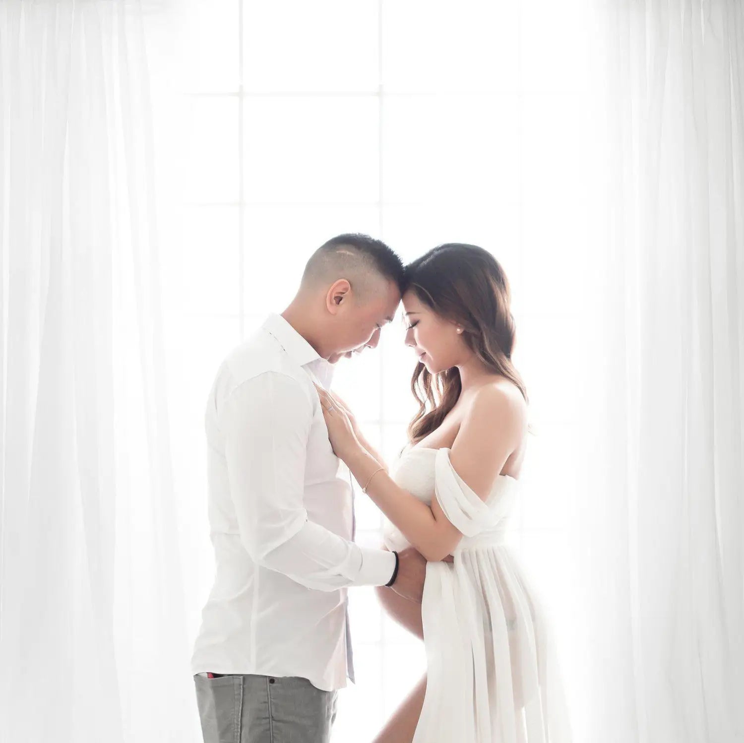 A pregnant woman in an off the shoulder white chiffon maternity gown stands tenderly with a man in a white shirt with soft light filtering through sheer curtains.