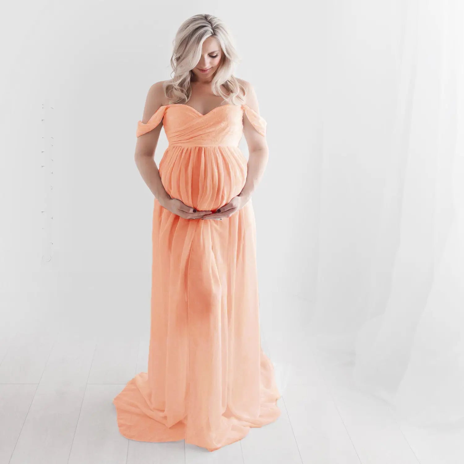 A flowing peach-colored chiffon maternity gown with an off the shoulder design and gathered waist for photoshoots.