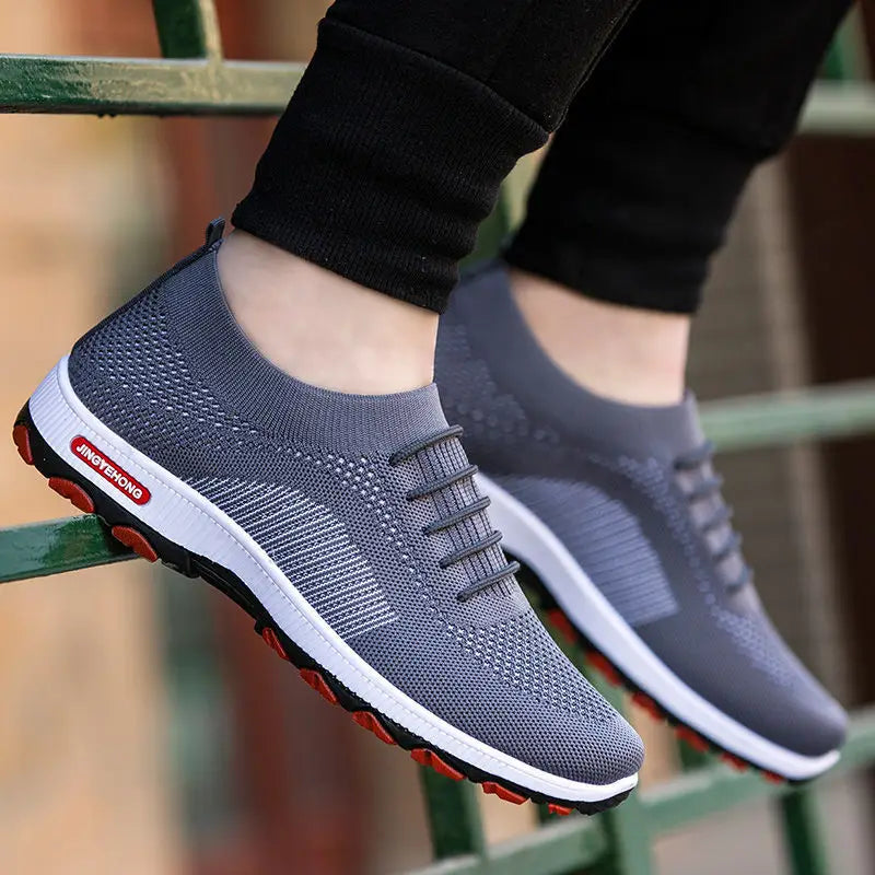 Herren-Strickatmungsaktive Freizeitsportschuhe in Grau mit weißen und roten Akzenten an der Sohle, mit atmungsaktivem Mesh-Obermaterial und schwarzen Schnürsenkeln.
