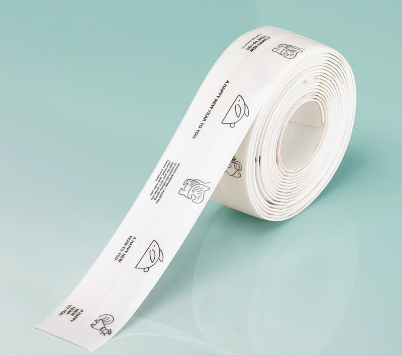 Un rouleau de ruban adhésif d’étanchéité pour cuisine en PVC blanc, imperméable et anti-moisissure, comportant des dessins au trait noirs et un texte incluant « Bonne année » à vos côtés, ainsi qu’un poisson et des personnages simples.