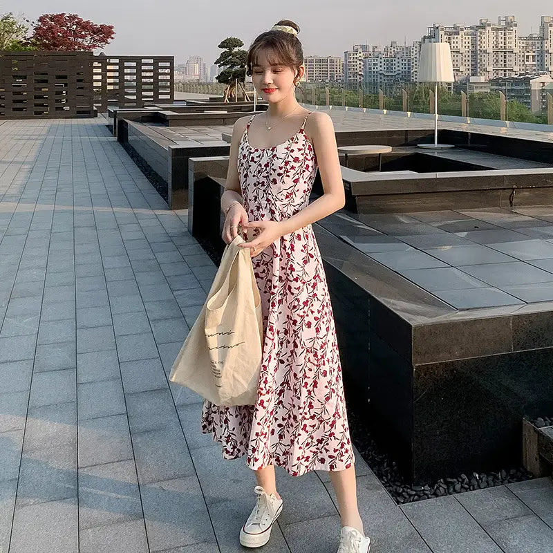 A woman wears a floral print halter midi dress for women featuring a flowing sleeveless design with delicate red and green floral patterns on a soft pink background.