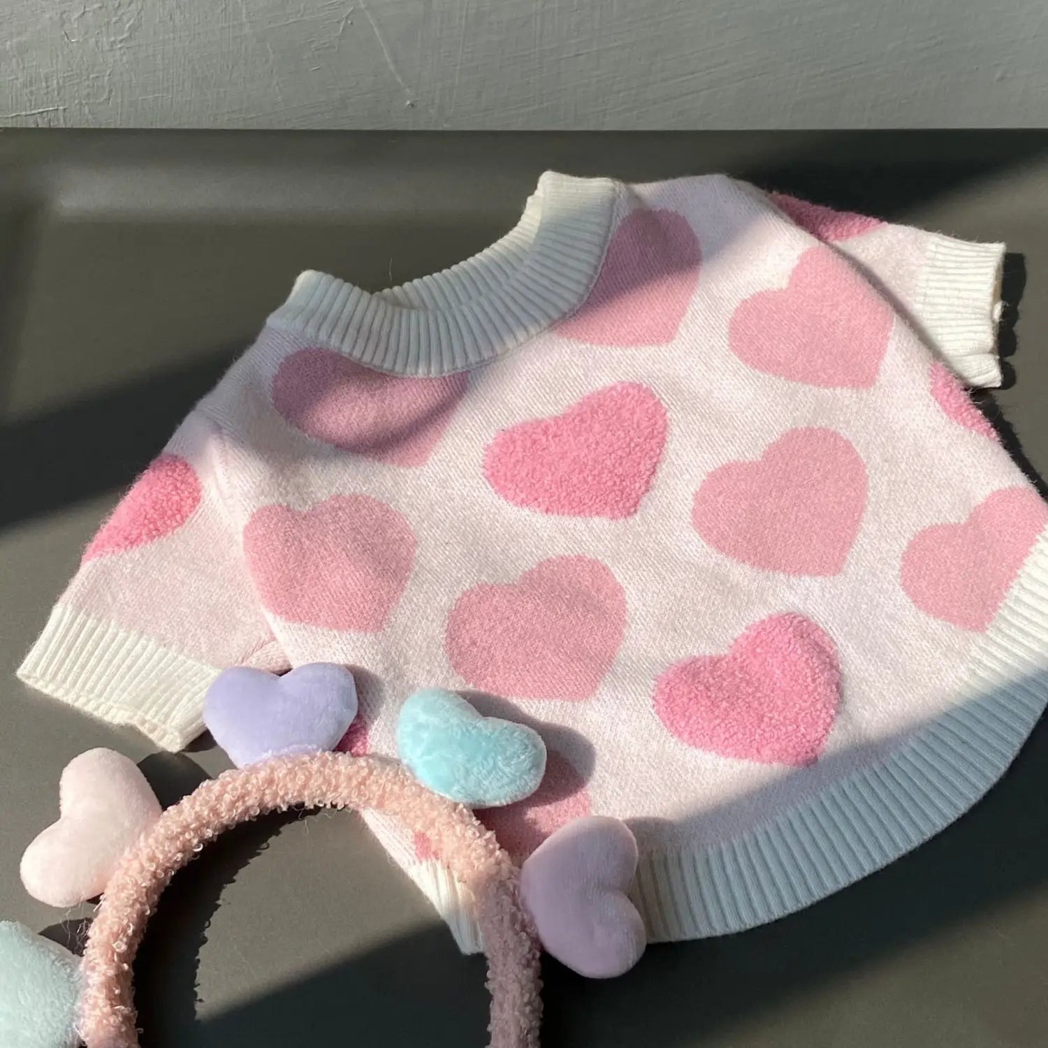 A soft pink and white heart-patterned knit dog sweater with fluffy pastel-colored heart-shaped accessories designed for winter pet apparel.