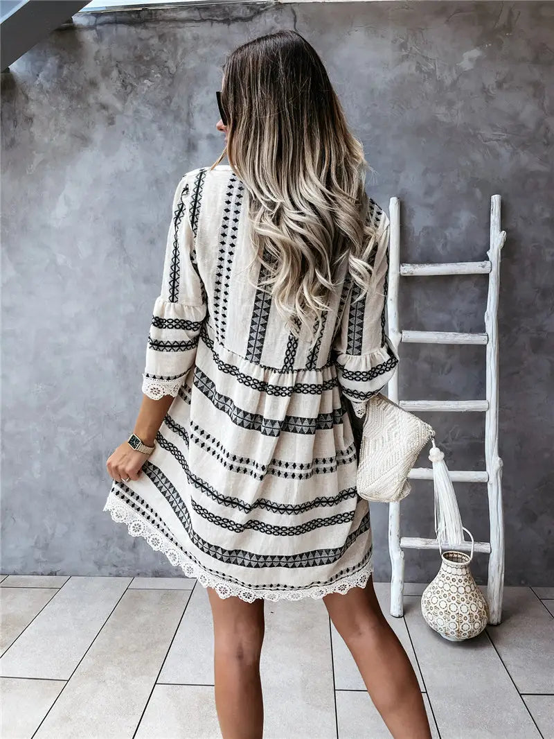 A woman wears a cream-colored boho geometric V-neck mini dress with black striped patterns and lace trim along the sleeves and hem.