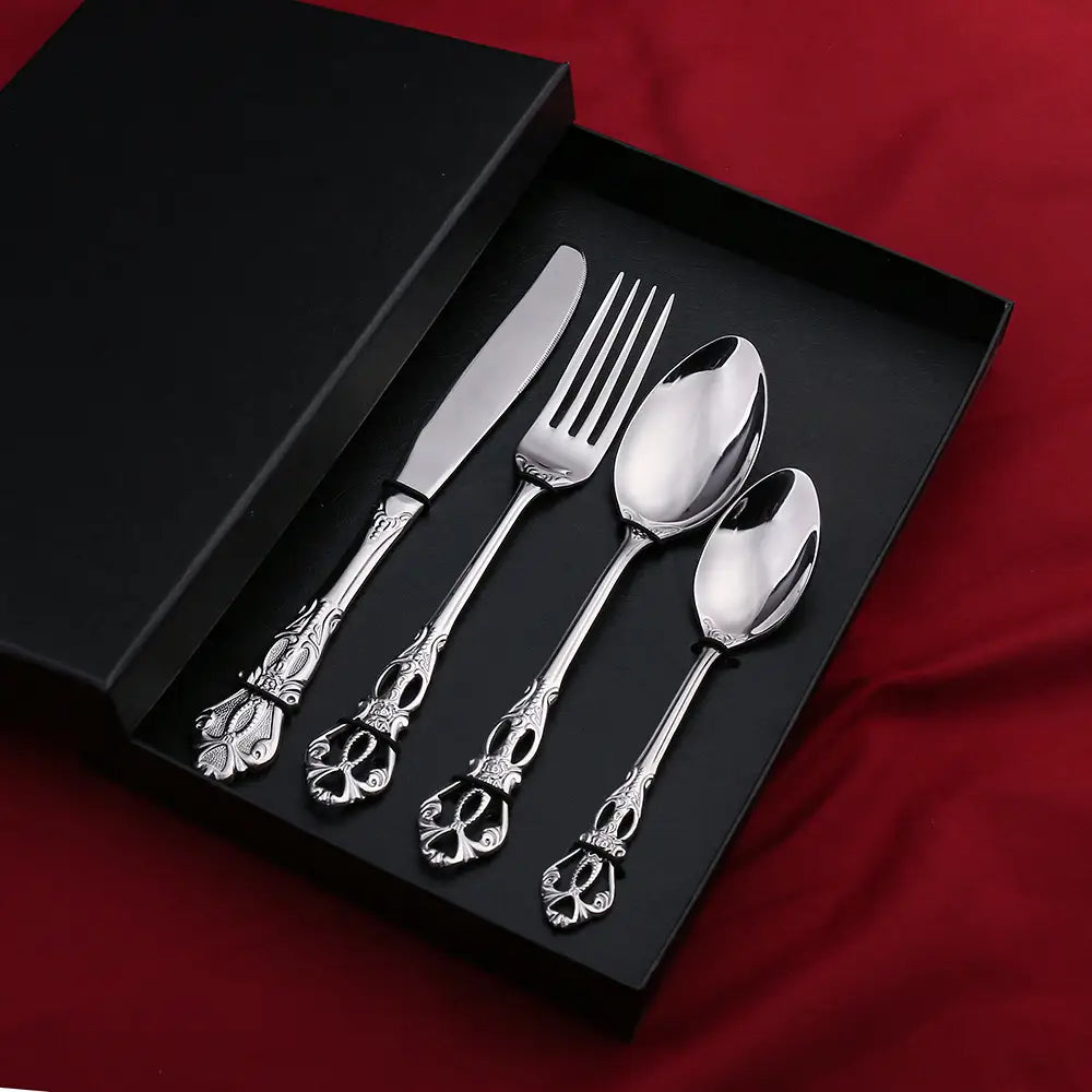 Un ensemble de couverts en acier inoxydable de la Cour Royale argenté, orné, avec des motifs de crânes sur les poignées, comprenant un couteau, une fourchette et deux cuillères, disposés avec soin dans une boîte-cadeau noire.