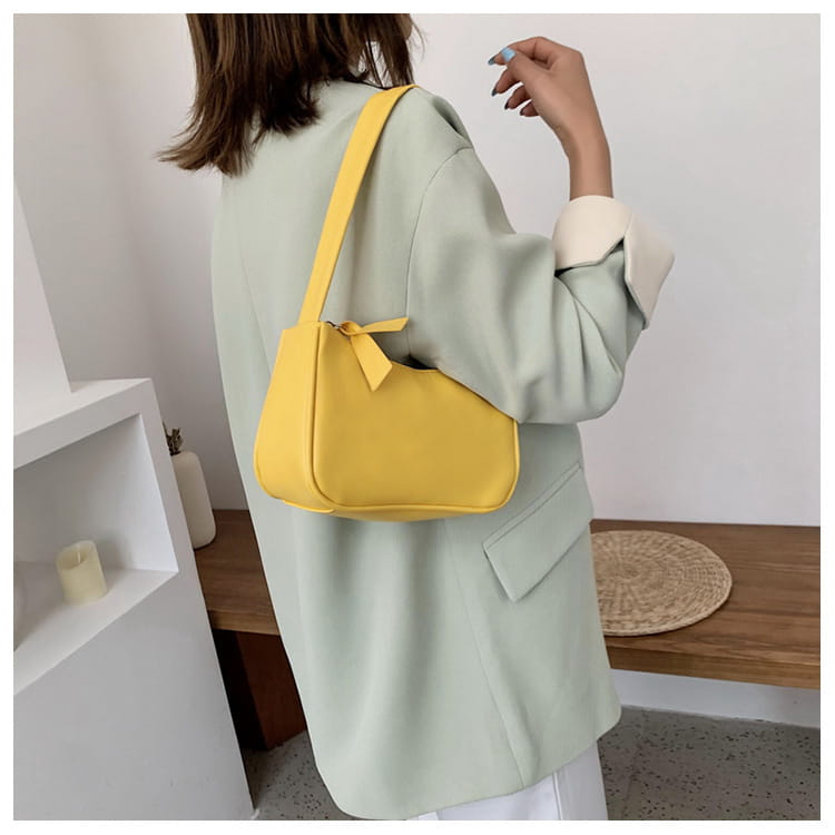 Eine minimalistische Umhängetasche aus Kunstleder mit gebogenem Unterarmgriff in leuchtendem Gelb, mit passendem Riemen und dekorativer Schleife am oberen Bereich.