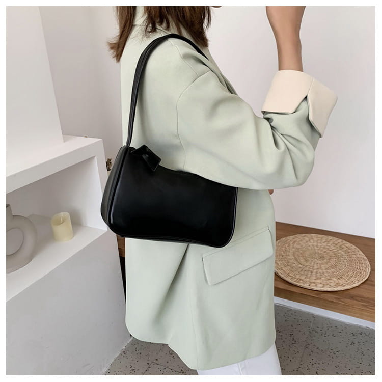 Eine schwarze minimalistische Umhängetasche aus Kunstleder mit gebogenem Unterarmgriff, mit einem eleganten Design und einer kleinen Schleifendetailierung im oberen Bereich.