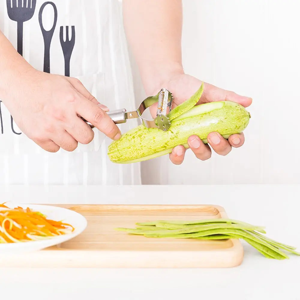 Silver stainless steel 2 in 1 potato peeler and julienne grater with a curved blade and ergonomic handle peeling a green cucumber