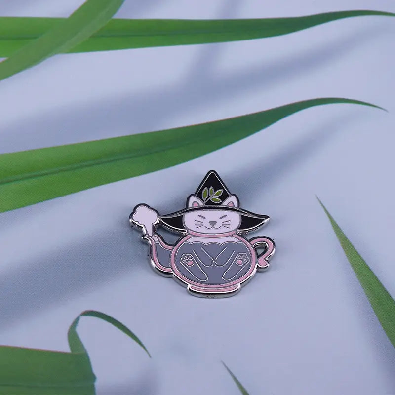 A charming copper enamel pin featuring a white cat wearing a black witchs hat with green leaves sitting inside a pink-rimmed teapot.