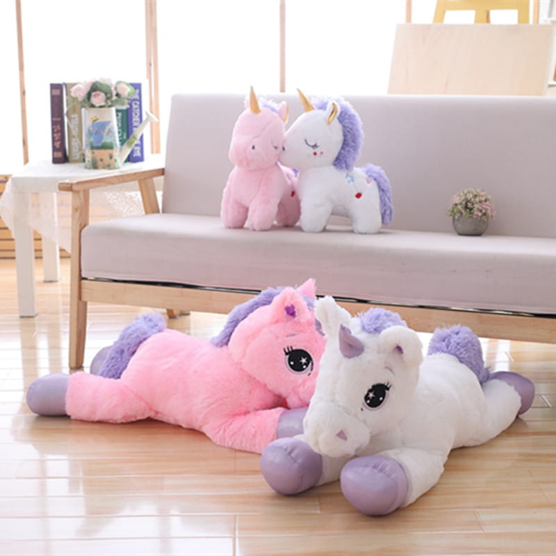 pink and white plush unicorn toys with purple manes tails and hooves lying on a wooden floor soft stuffed animals for children