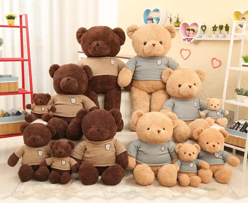 Peluches de osos de peluche en varios tamaños con pelaje marrón claro y beige, con suéteres de punto color camel o gris, que presentan un pequeño emblema con forma de escudo en el pecho.