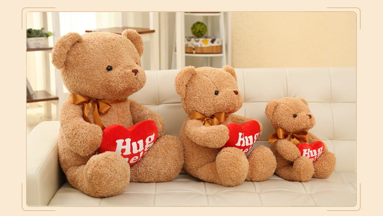 Tres osos de peluche de tono beige claro, cada uno con un lazo dorado y sosteniendo un corazón rojo con la palabra «abrázame», dispuestos en tamaños decrecientes sobre un sofá blanco, como peluches de osos con suéteres de punto de 55 cm