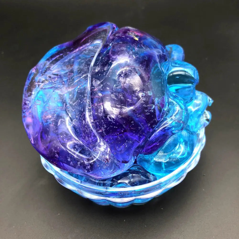 A translucent blue and purple glass sculpture of a rose with intricate petal details resting in a matching scalloped glass bowl featured in the gradient crystal mud slime blue and purple putty toy product.