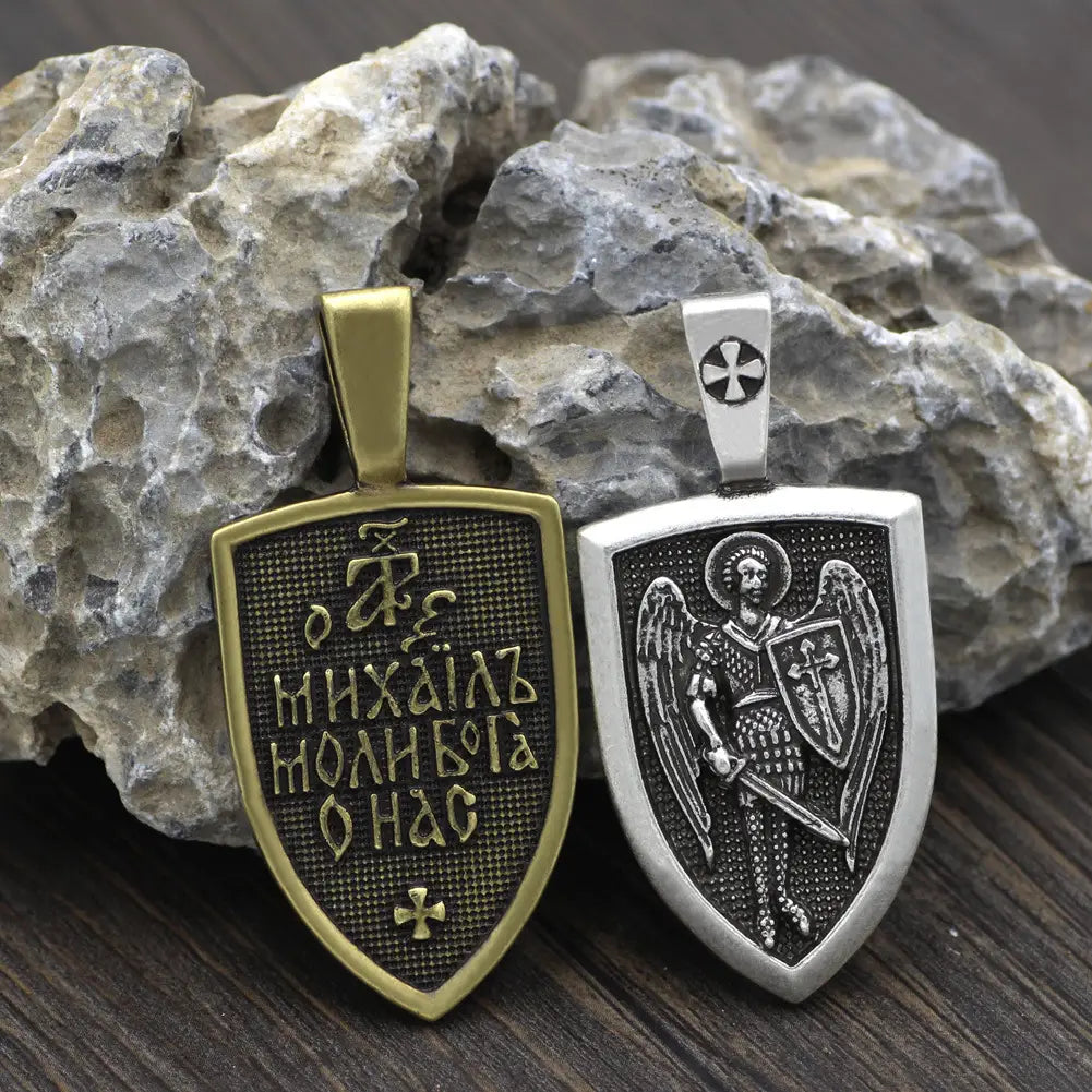 Two shield-shaped metal pendants from Saint Michael Archangel shield pendant on braided cord necklace featuring a gold-tone engraved cyrillic script and a silver-tone detailed angelic figure holding a sword and shield.