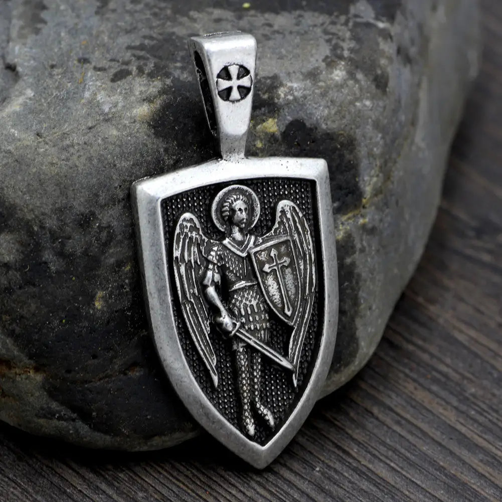 Silver shield-shaped pendant featuring an intricately engraved Saint Michael Archangel in armor holding a sword and shield with detailed wings and halos on a braided cord necklace.