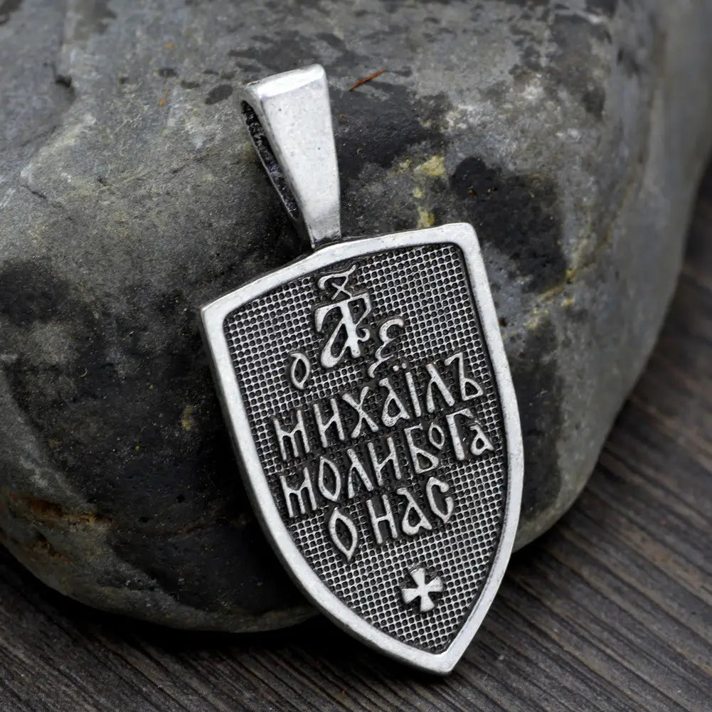 Silver shield-shaped pendant in textured grid pattern with engraved cyrillic script and small cross Saint Michael Archangel shield pendant on braided cord necklace.