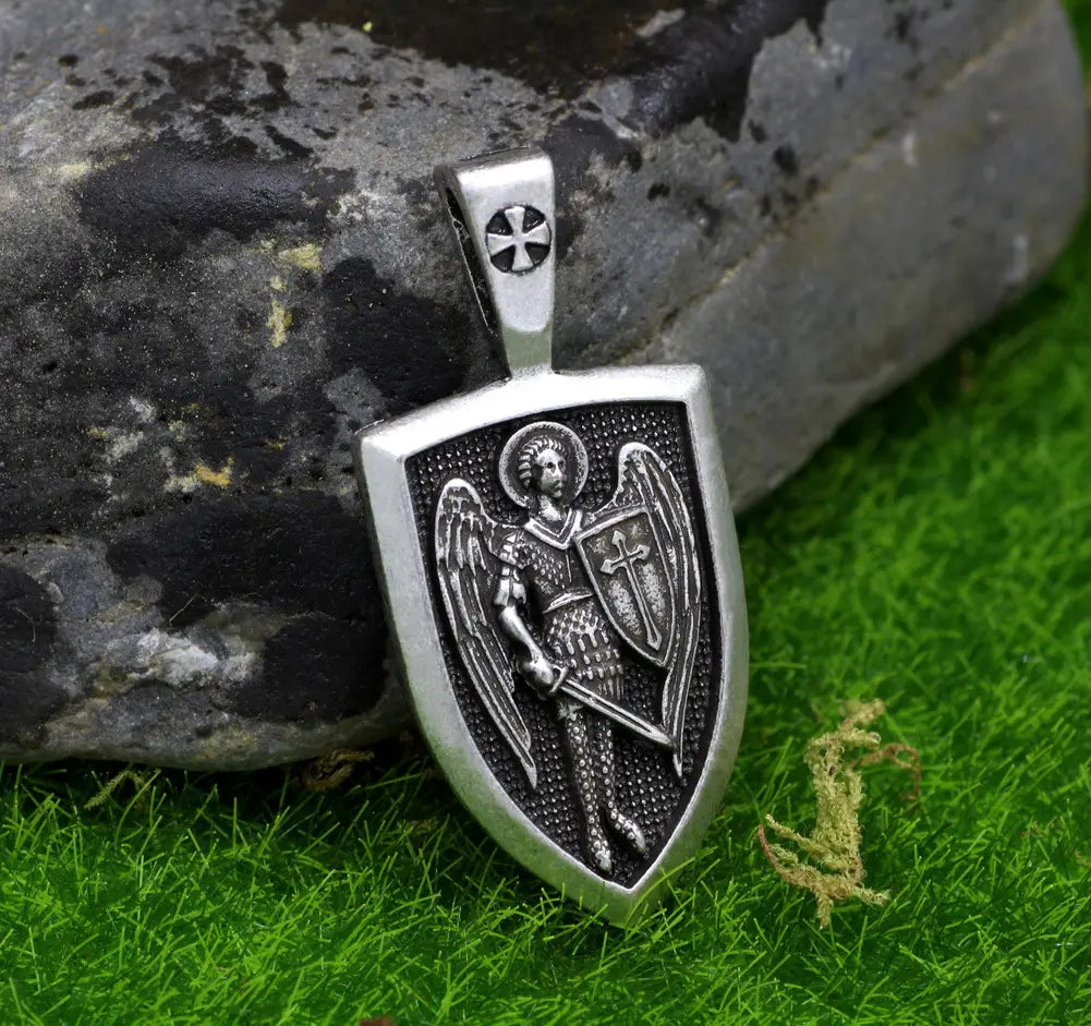 Silver shield-shaped pendant featuring an intricately engraved Saint Michael Archangel in armor holding a sword and shield with detailed wings and a cross emblem on a braided cord necklace.