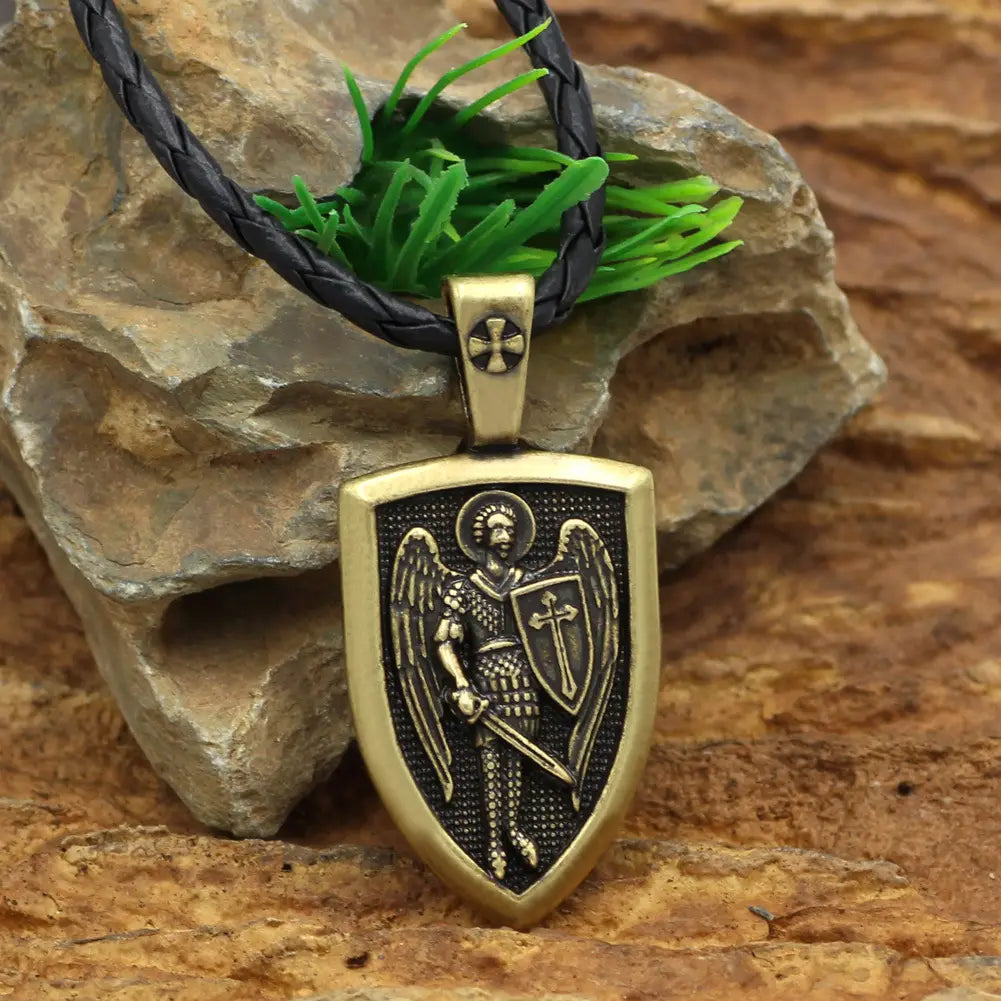 Antique brass shield-shaped Saint Michael Archangel pendant featuring a detailed angel in armor holding a sword and shield, suspended from a black braided cord necklace.