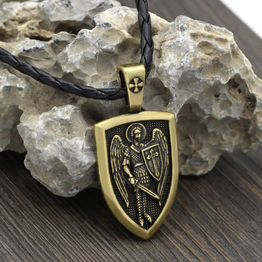 Gold-toned shield-shaped pendant featuring an engraved Saint Michael archangel in armor holding a sword and shield, suspended from a black braided cord necklace.