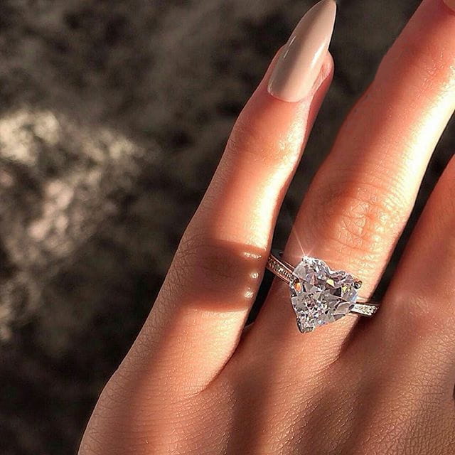 A sparkling heart shaped zircon engagement ring with white gold plating on a finger with long beige nails.